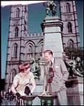Tourists at water fountain in the Place d'armes, Montreal. In background is the Maisonneuve monument at Notre Dame Church. [Touristes à une fontaine d'eau au Place d'armes, Montréal. À l'arrière-plan est le monument Maisonneuve à la première église de Ville-Marie.] juin 1950.
