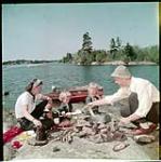 Family making a shore dinner on Stony Lake. Mother, Father, and two little girls around fire in the foreground. août 1951.
