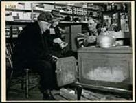 G.W. Bourbonnais, à droite, se repose dans sa chaise à bras, près du poêle à bois dans son magasin général, et converse avec deux clients,  à Fort-Coulonge n.d.