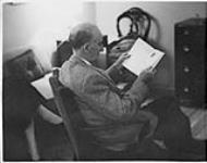  Wilson P. MacDonald sitting in a chair, reading [1955]