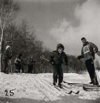 [Young boy with his hood up and smiling, sets off down a hill skiing as a male instructor watches] [between 1953-1964]