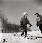 [Young girl wearing light coloured, fluffy winter hat, sets off down a hill skiing as a male instructor watches] [between 1953-1964]