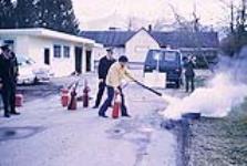 Fire marshal instructing man how to use a fire extinguisher to put out a tire fire February 1972