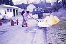 Commissaire des incendies montrant à un homme comment utiliser un extincteur pour éteindre un pneu en feu février 1972