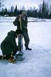 Woman lowering a bucket into a hole in the ice, man with a camera around his neck stands over her watching February 1962