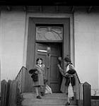 Two girls stand in front of their house with Anne Piper at the door [ca1954-1963]