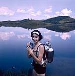 Close-up portrait of female scuba diver Heather McEwen at water's edge wearing oxygen tanks on her back and holding diving a mask in her hands 1954