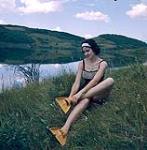 Female scuba diver Heather McEwen seated on the grass at water's edge putting on yellow flippers 1954