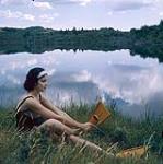 Female scuba diver Heather McEwen seated on the grass at water's edge putting on yellow flippers 1954