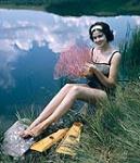 Female scuba diver Heather McEwen seated on the grass at water's edge holding a pink fan coral 1954