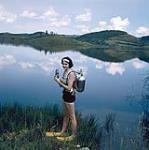 Female scuba diver Heather McEwen standing at water's edge wearing oxygen tanks on her back, yellow flippers, and holding a diving mask in her hands 1954
