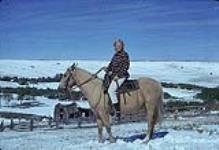 Une femme assis sur un cheval en  hiver; on voit une grange derrière la colline à l'arrière-plan. Flat Creek ou Timber Ridge September, 1962