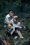 Homme âgé portant un imperméable transparent, assis dans le bois et mangeant August, 1963