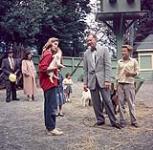 Woman wearing red shirt holding a piglet. Man and teenage boy look on. Children's Zoo, Stanley Park 1954