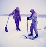 Two men making a hole in the ice for ice fishing, Patuanak, Saskatchewan [Pierre Lariviere is on the right and the man on the left is probably George Campbell or Alec Campbell] mars 1955