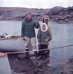 Blanc, Inuit et garçon inuit debout au bord de l'eau, près d'un canot. Arctique/Nord canadien [between June 17-October 31, 1960].