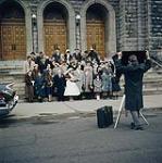 A formal portrait of a wedding. The wedding party is standing out in front of the Church. In the foreground can be seen the back of a photographer taking the wedding portrait. Montréal, Québec 1959