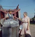 A dairy farmer sits on a tractor, facing the camera. His wife stands beside him. 1957