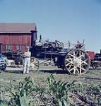 A farmer stands beside an old steam engine which was used for cultivating the farmlands in bygone eras. In the foreground is some young corn. août 1959
