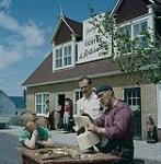 Tourists watching Andre Bourgault at work in front of his Centre D'Artisanat, St. Jean Port Joli, P.Q. June 1959