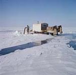 Horses pulling wagon behind man holding fishing net in hole in the ice. Lake Manitoba. [Homme tenant un filet de pêche près d'un trou dans la glace. Derrière, des chevaux tirant une voiture. Lac Manitoba.] 1961