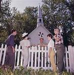 Two young men and two young women speaking with man over picket fence outside church. Bagotville, Quebec. July 1961