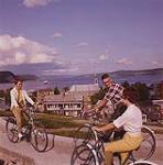 A young man bicycling along path with two young women.  July 1961
