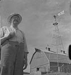Saskatoon et blé, fermier debout à l'extérieur d'une grange [between 1939-1951].