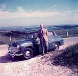 Lionel Gilliat standing beside a blue car [ca 1953-1964]