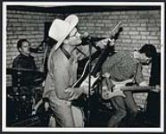 Marshall Crenshaw portant un chapeau de cow-boy, jouant de la guitare et chantant au micro sur scène octobre 1987