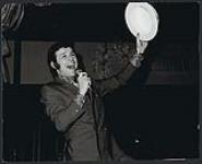 Unidentified musician singing into a microphone, holding a hat in the air [entre 1968-1975].