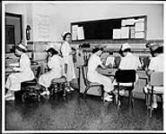 Five nurses sit doing paper work at desks while one stands and looks on. Northern Ontario or Manitoba (?). Nurses and Nursing. Department of Citizenship and Immigration, Information Division 1956