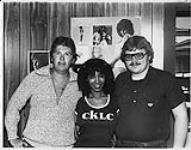 Portrait of three people, two women and one man. Woman in the middle wears a CKLC t-shirt. Barbara Streisand poster in the background [entre 1977-1980].