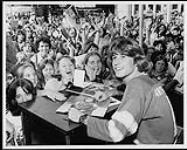 Andy Gibb sitting at a desk and signing albums, in front of a crowd of young women [between 1977-1980].