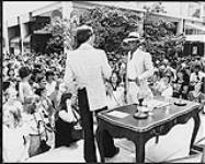 Paul Anka entouré de fans sur scène qui s'entretient avec un autre homme. Les deux hommes tiennent un microphone [between 1975-1980].
