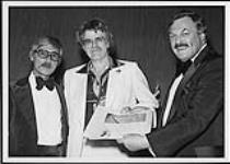 Dallas Harms (centre) standing with two men in tuxedoes, holding a certificate [entre 1975-1980].