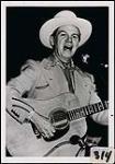 Earl Heywood wearing a cowboy hat, playing a guitar and singing into a microphone [between 1985-1990].