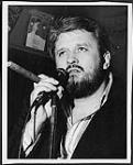 Ronnie Hawkins smoking a cigar and holding a microphone [between 1970-1975].