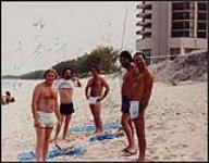 Johnny Burke and members of Eastwind standing on a beach in swimsuits [between 1975-1980].