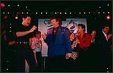 Bobby Curtola souriant, tandis qu'un imitateur d'Elvis Presley chante devant lui [between 1988-1995].