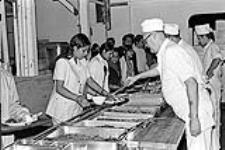 Caption: "Dinner is served! The Ugandan Asians arriving at CFB Montreal Longue Pointe Garrison receive specially prepared Indian cuisine. The CAF cooks received professionnal [sic] instructions on the correct way to prepare the many different types of curried dishes."