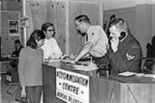 Caption: "Mr. and Mrs. Anil Lalani (left) receive accommodation instructions from Sgt Bill Somers (centre) while Cpl Robert Boudreau makes arrangements from the phone."