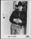 Photo publicitaire de Ian Tyson portant un chapeau de cow-boy et des gants de travail, appuyé sur une porte de bois [entre 1983-1984].