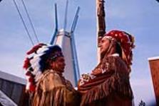 Indians of Canada pavilion - two men wearing headdresses in front of totem pole [1963-1967]