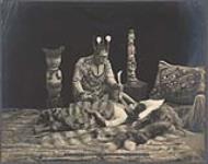 [Studio portrait of posed scene of Da-Yuc-Hene, Tlingit healer, leaning over woman lying down wrapped in fur blankets]. Original title Indian Witch Doctor or Sha-man, healing a sick woman 1906
