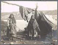 [Two Gwich'in women at their camp with a tent and large hide (caribou or moose) hanging to dry] [between 1889-1942]