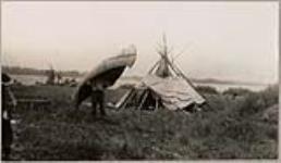 [Man carrying a birchbark canoe away from the shore and a woman seated inside a canvas tent] [ca. 1916]