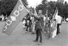 Full profile view of man in camouflage jacket holding a Haudenosaunee Confederacy flag with more flags and crowd behind him on golf course 11 July 1995