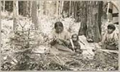 [Anishinaabe woman with two children splitting spruce root for canoe making] 1919