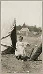 [Young Anishinaabe girl, wearing ankle-wrap moccasins, standing outside a tent] 1920